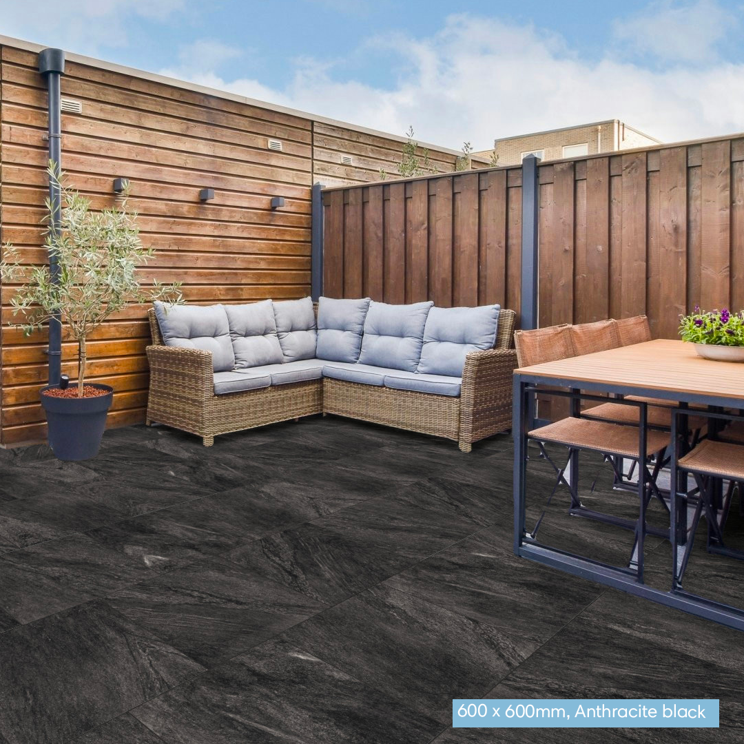 Outdoor patio area with anthracite black stone pavers, a wicker sectional sofa with light blue cushions, a wooden dining table, and a tall wooden fence under a blue sky.