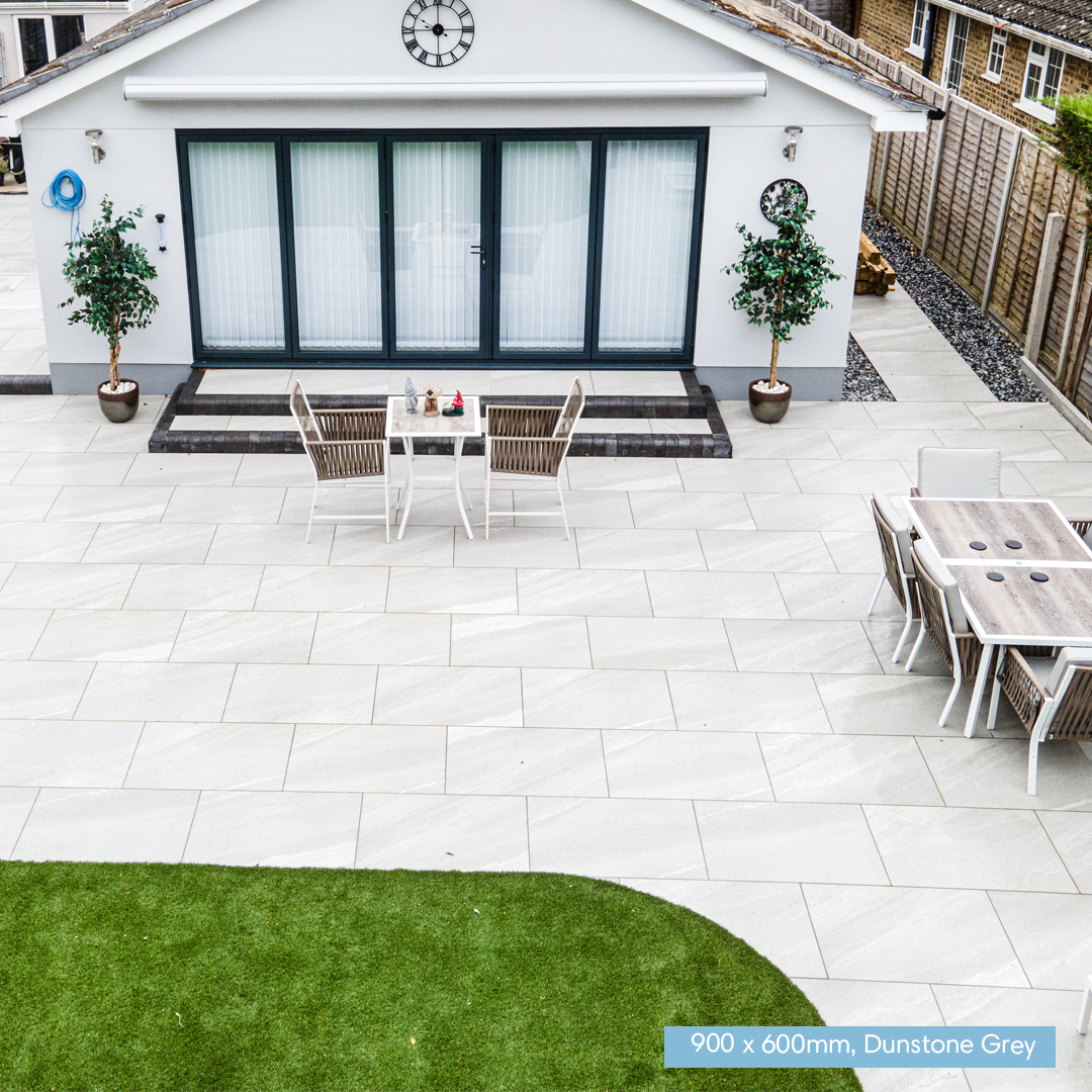 A high-angle view of a modern outdoor patio featuring large, light grey stone tiles. In the background is a white detached garage with black-framed glass doors, flanked by potted trees.