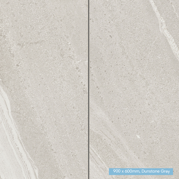 Two large stone-look tiles in a light warm gray finish with subtle natural veining, separated by a thin dark grout line. A small blue label in the bottom right reads '900 x 600mm, Dunstone Grey'.